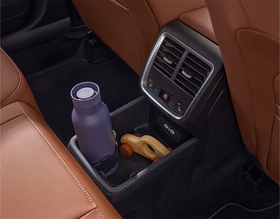 Eine Nahaufnahme zeigt die Rückseite der Mittelkonsole eines Škoda Octavia mit der Design Selection Suite in Cognac. Unter den Lüftungsdüsen und den Bedienelementen für die hintere Klimatisierung befinden sich zwei USB-C-Anschlüsse und ein Ablagefach. In diesem stehen eine lila Trinkflasche und ein Holzauto.