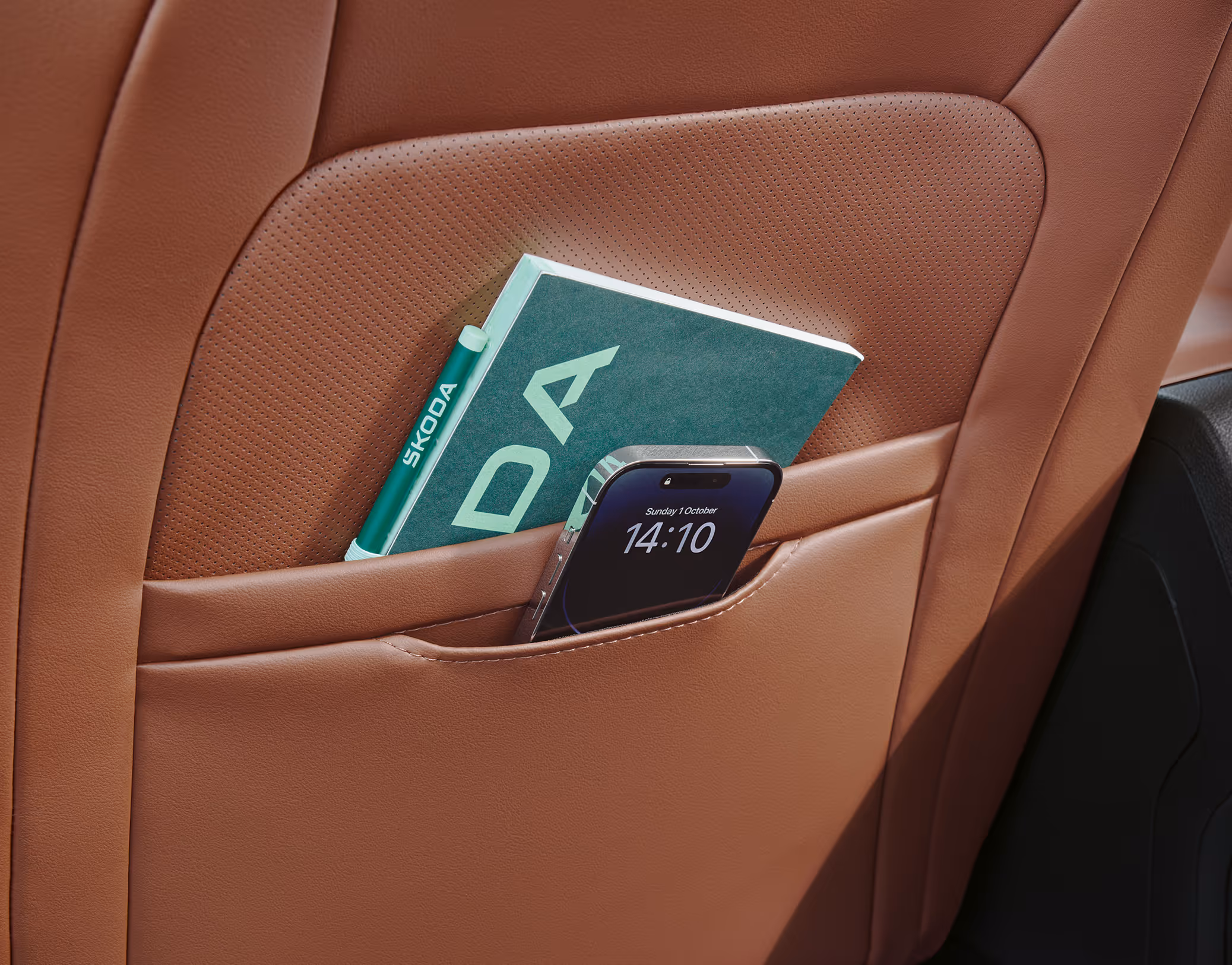 Eine Nahaufnahme zeigt die Ablagetaschen an der Rückseite eines Vordersitzes im Škoda Octavia. In den Taschen des cognac-farbenen Ledersitzes der Design Selection Suite stecken ein Smartphone, dessen Display die Uhrzeit anzeigt, und ein Notizbuch.