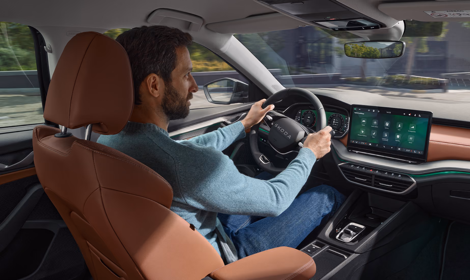 Eine Ansicht aus dem Innenraum eines Škoda Octavia zeigt einen Mann, der das Fahrzeug fährt. Das Interieur der Design Selection Suite in Cognac mit seinen cognac-farbenen Ledersitzen und Armaturenbrett-Applikationen wird durch eine grüne Ambientebeleuchtung hervorgehoben. Der Fahrer hat beide Hände am Lenkrad, während das digitale Cockpit und das Infotainment-Display eingeschaltet sind.