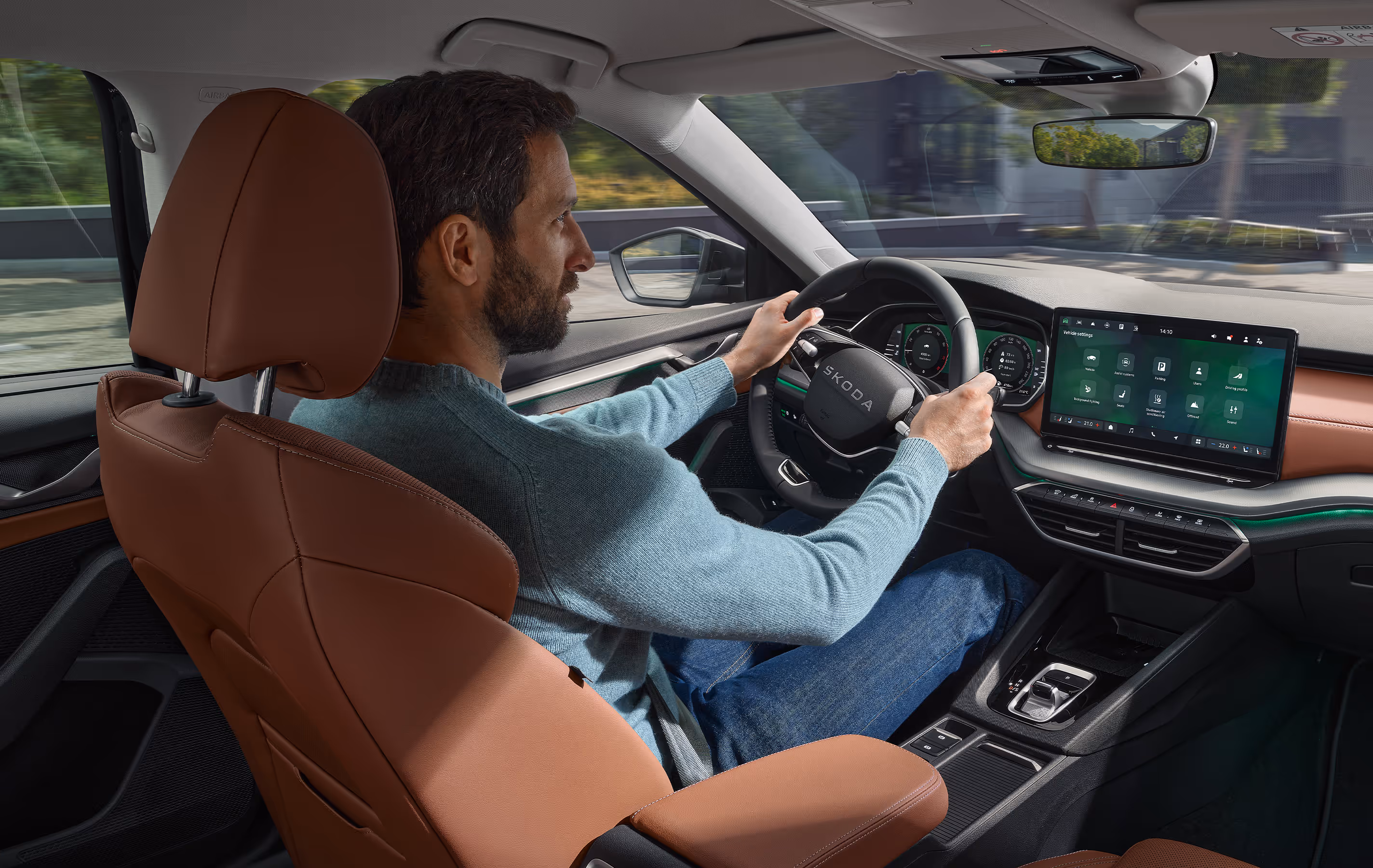 Eine Ansicht aus dem Innenraum eines Škoda Octavia zeigt einen Mann, der das Fahrzeug fährt. Das Interieur der Design Selection Suite in Cognac mit seinen cognac-farbenen Ledersitzen und Armaturenbrett-Applikationen wird durch eine grüne Ambientebeleuchtung hervorgehoben. Der Fahrer hat beide Hände am Lenkrad, während das digitale Cockpit und das Infotainment-Display eingeschaltet sind.