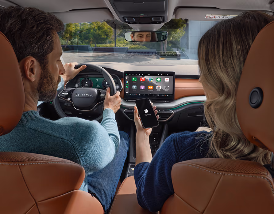 Eine Ansicht aus dem Innenraum eines Škoda Octavia zeigt die Nutzung der Wireless SmartLink Funktion. Während ein Mann am Steuer sitzt, hält die Beifahrerin ein Smartphone mit dem CarPlay-Logo, dessen Benutzeroberfläche auf dem großen, freistehenden Infotainmentdisplay gespiegelt wird. Die Szene zeigt das Interieur der Design Selection Suite in Cognac, während durch die Windschutzscheibe eine begrünte Umgebung sichtbar ist.