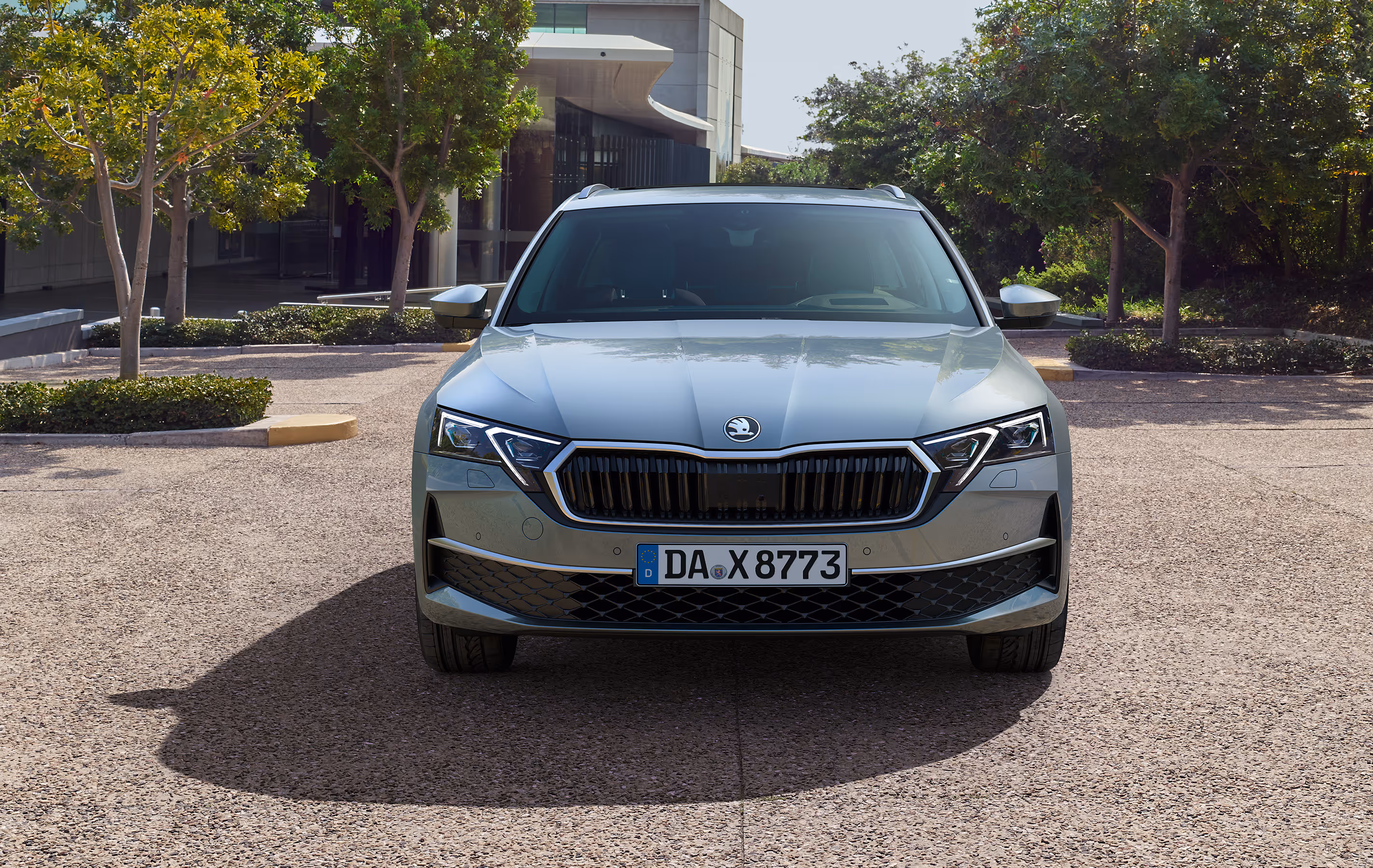 Ein Škoda Octavia Combi in der Lackierung Smokey Diamond-Silber Metallic ist frontal auf einem gepflasterten Platz geparkt. Im Hintergrund sind Bäume und ein modernes Gebäude zu sehen.