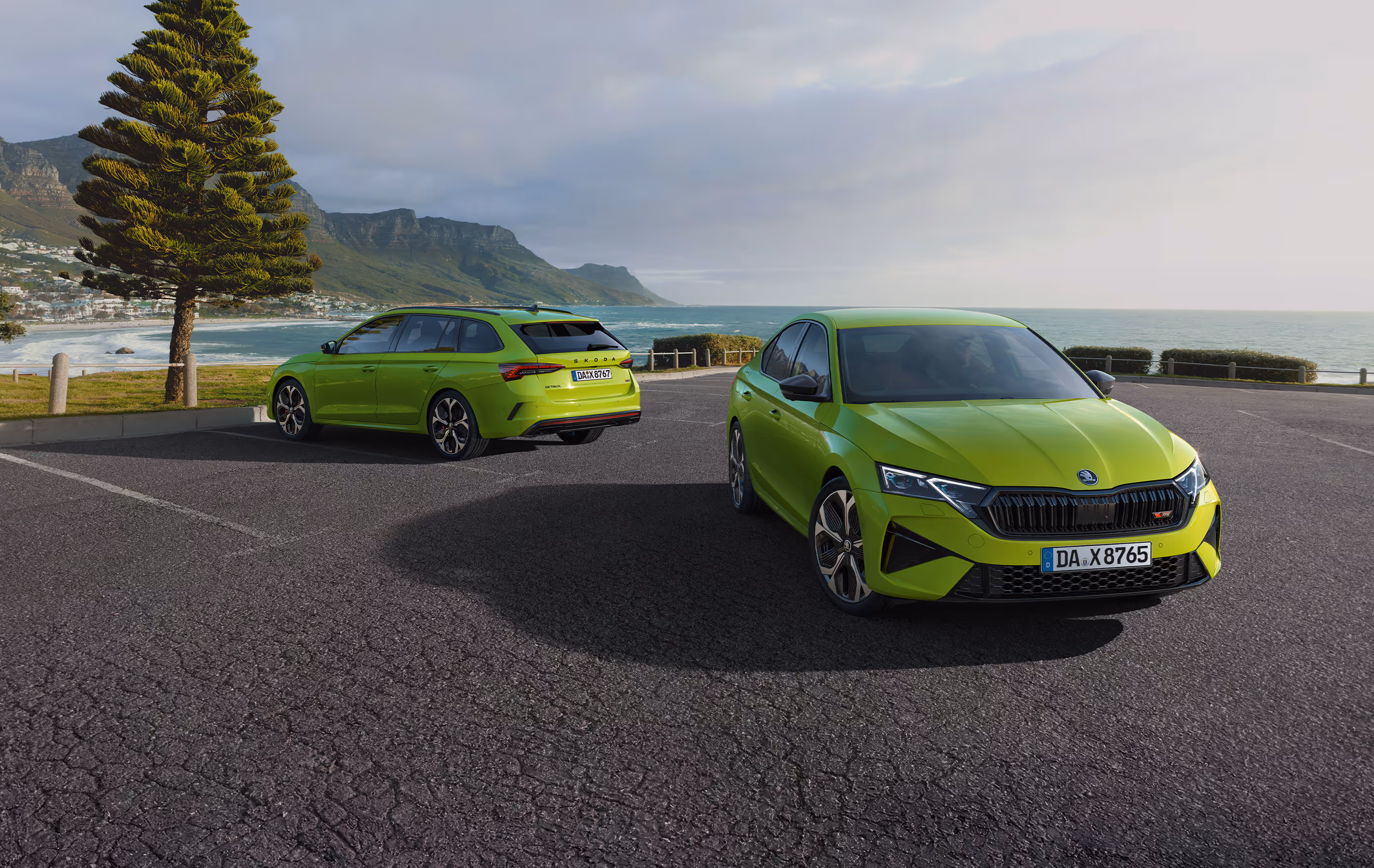 Zwei Škoda Octavia RS Modelle in der Lackierung Mamba-Grün sind auf einem Parkplatz an einer Küste geparkt. Die Limousine steht im Vordergrund in einer Dreiviertelansicht von vorne, während der Combi dahinter von schräg hinten zu sehen ist. Im Hintergrund erstreckt sich eine Bucht mit Strand und eine bergige Küstenlinie.