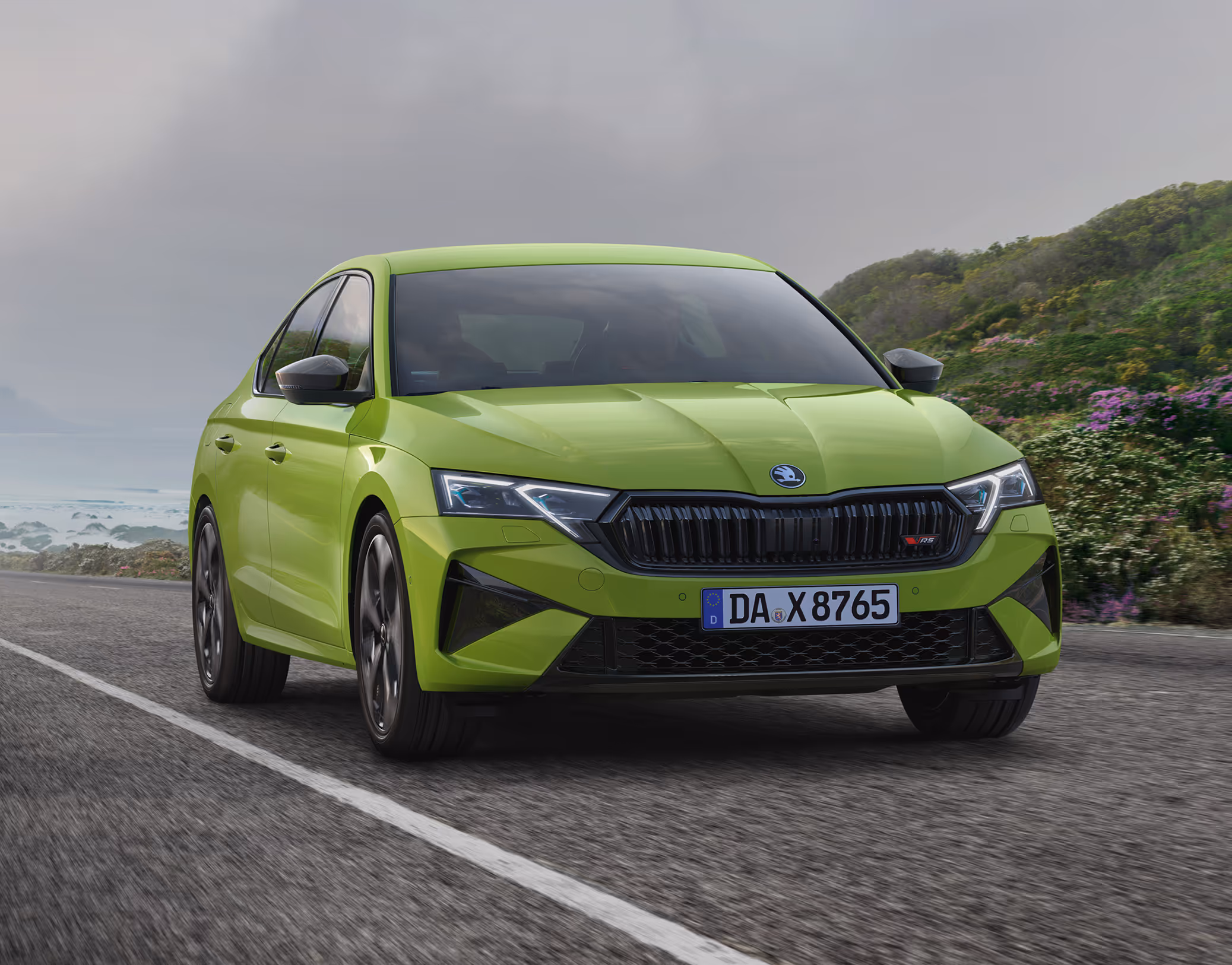 Ein Škoda Octavia RS Limousine in der Lackierung Mamba-Grün fährt auf einer Küstenstraße. Das Fahrzeug wird in einer dynamischen Dreiviertelansicht von vorne gezeigt, die die sportlichen Designelemente wie den schwarzen Kühlergrill hervorhebt. Im Hintergrund ist das Meer und eine bewachsene, felsige Küste unter einem bewölkten Himmel zu sehen.