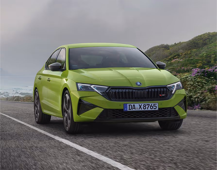 Ein Škoda Octavia RS Limousine in der Lackierung Mamba-Grün fährt auf einer Küstenstraße. Das Fahrzeug wird in einer dynamischen Dreiviertelansicht von vorne gezeigt, die die sportlichen Designelemente wie den schwarzen Kühlergrill hervorhebt. Im Hintergrund ist das Meer und eine bewachsene, felsige Küste unter einem bewölkten Himmel zu sehen.