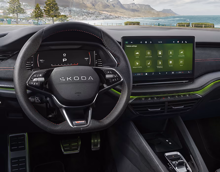 Eine Ansicht aus dem Cockpit eines Škoda Octavia RS zeigt das sportliche Interieur. Das perforierte Sport-Multifunktionslenkrad mit RS-Plakette, die Pedalerie in Edelstahloptik und die grüne Ambientebeleuchtung prägen den Innenraum. Durch die Windschutzscheibe ist eine malerische Küstenlandschaft mit Bergen und Meer zu sehen.