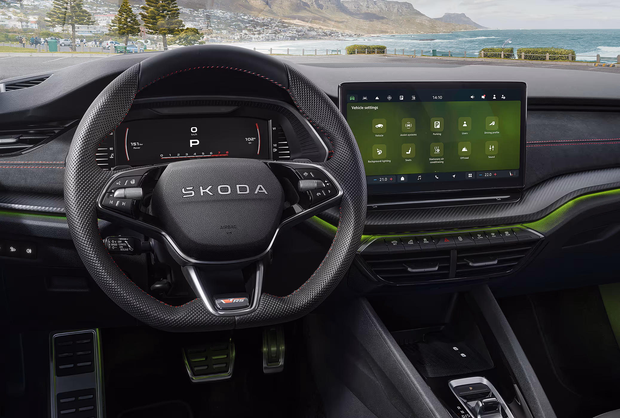 Eine Ansicht aus dem Cockpit eines Škoda Octavia RS zeigt das sportliche Interieur. Das perforierte Sport-Multifunktionslenkrad mit RS-Plakette, die Pedalerie in Edelstahloptik und die grüne Ambientebeleuchtung prägen den Innenraum. Durch die Windschutzscheibe ist eine malerische Küstenlandschaft mit Bergen und Meer zu sehen.