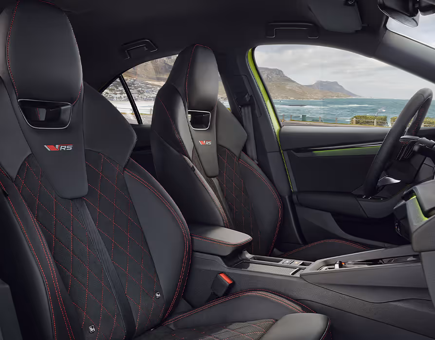 Eine seitliche Innenansicht des Škoda Octavia RS zeigt das sportliche Interieur. Die schwarzen Sportsitze mit integrierten Kopfstützen sind mit einer Rautensteppung und roten Kontrastnähten versehen, und das RS-Logo ist in die Kopfstützen eingestickt. Durch die Fenster ist eine Küstenlandschaft zu sehen.