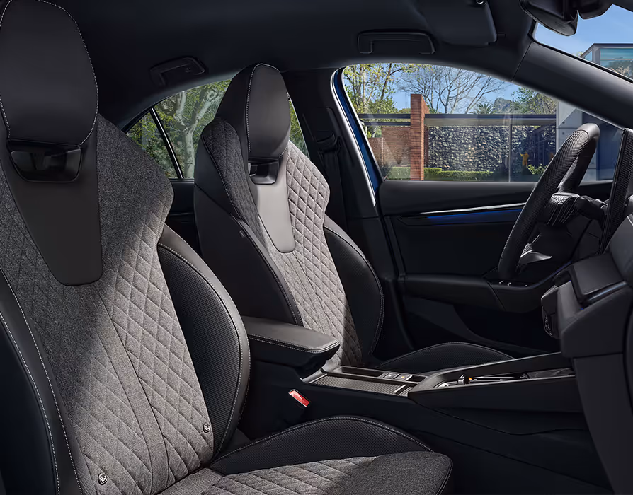 Eine seitliche Innenansicht des Škoda Octavia Sportline zeigt das sportliche Interieur der Design Selection Sportline. Die schwarzen Sportsitze mit integrierten Kopfstützen sind mit grauem, rautengestepptem Stoff und grauen Kontrastnähten versehen. Durch die Fenster ist eine begrünte, sonnige Außenanlage zu erkennen.