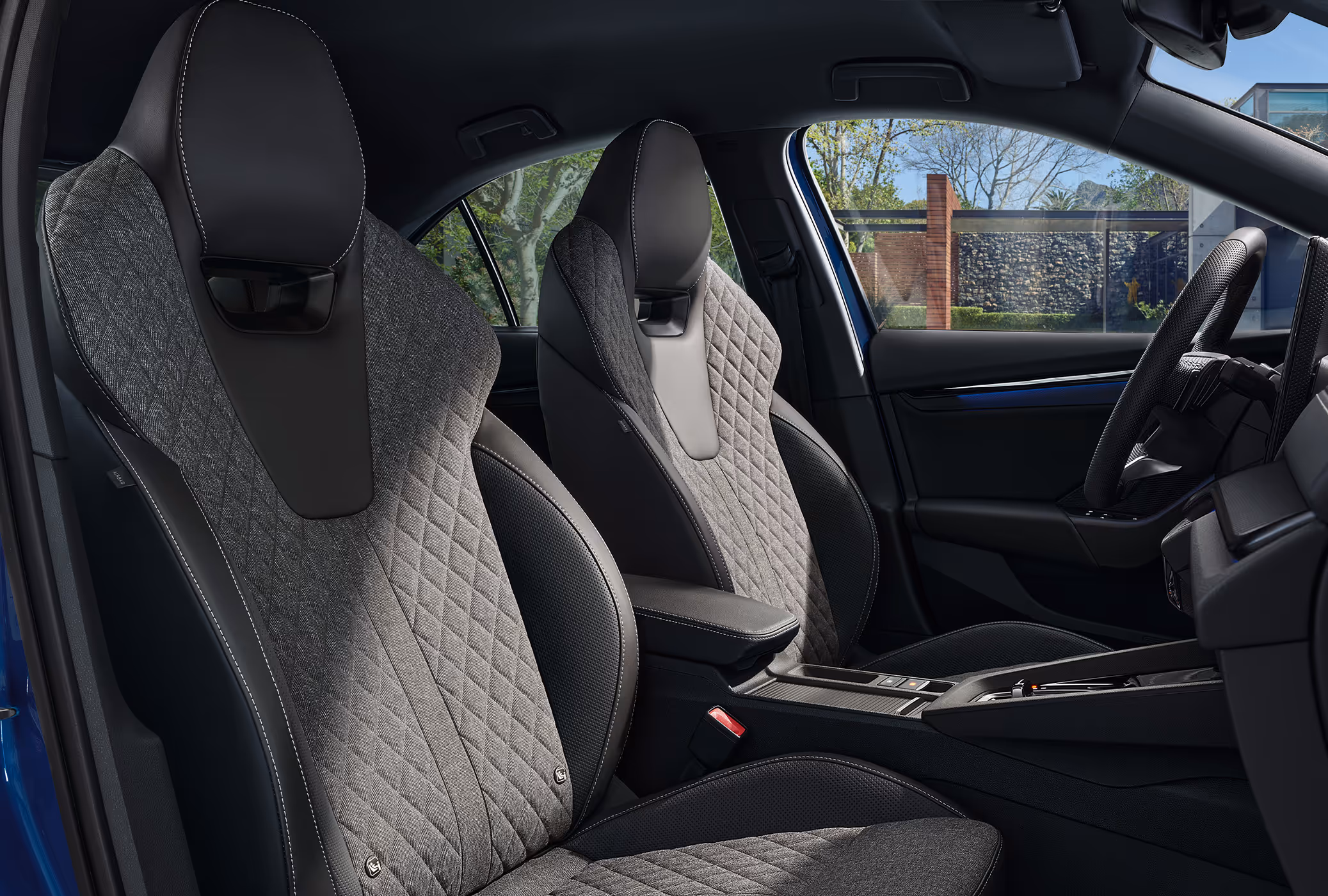 Eine seitliche Innenansicht des Škoda Octavia Sportline zeigt das sportliche Interieur der Design Selection Sportline. Die schwarzen Sportsitze mit integrierten Kopfstützen sind mit grauem, rautengestepptem Stoff und grauen Kontrastnähten versehen. Durch die Fenster ist eine begrünte, sonnige Außenanlage zu erkennen.