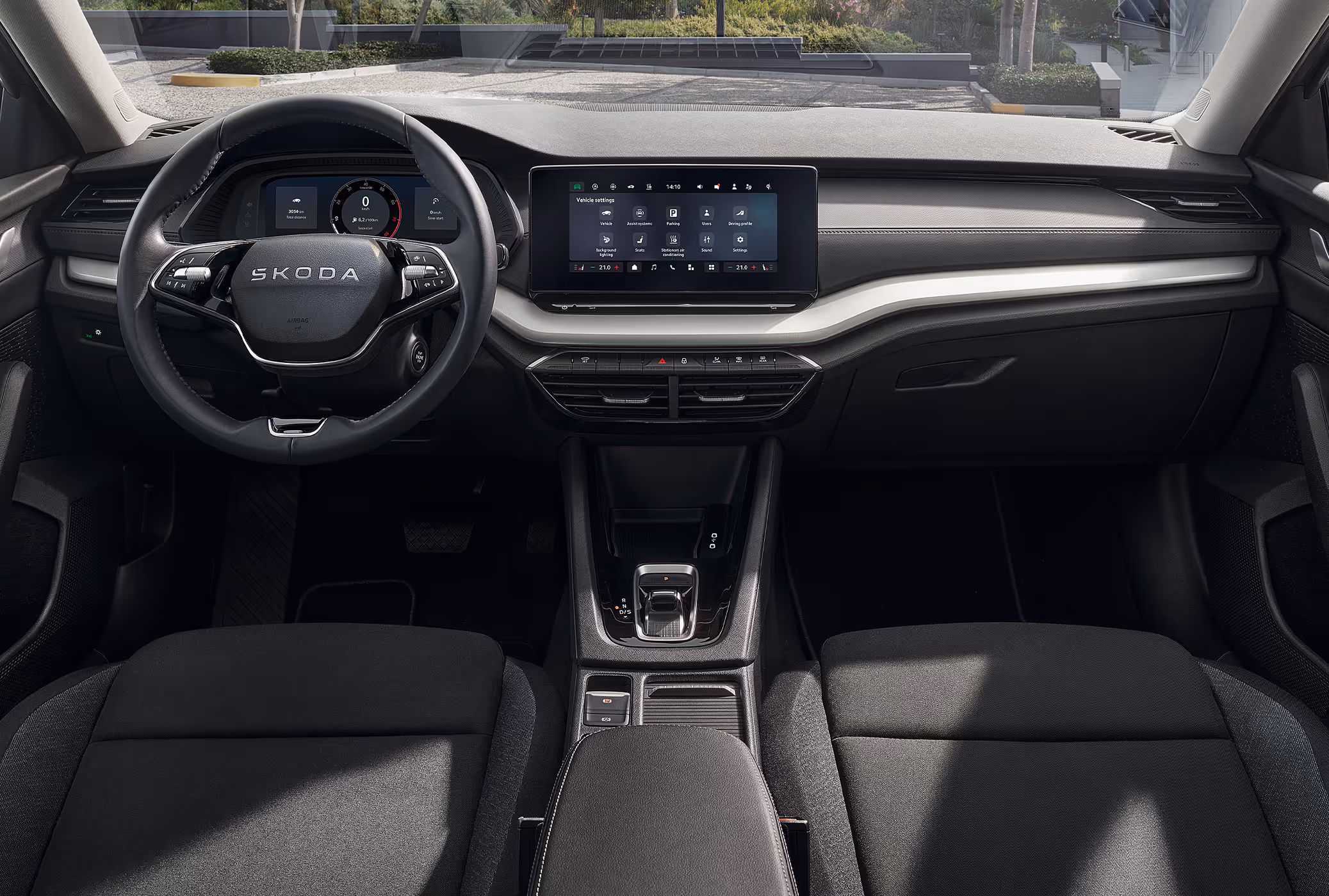 Eine weite Innenansicht zeigt das gesamte Cockpit eines Škoda Octavia mit der Ausstattungslinie Essence. Das dunkle Interieur ist mit schwarzen Stoffsitzen und einer silbernen Dekorleiste am Armaturenbrett ausgestattet. Durch die Windschutzscheibe ist eine begrünte, städtische Umgebung zu sehen.