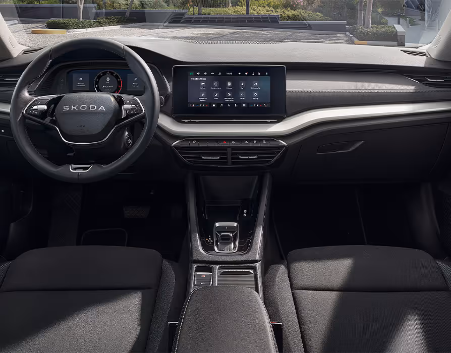 Eine weite Innenansicht zeigt das gesamte Cockpit eines Škoda Octavia mit der Ausstattungslinie Essence. Das dunkle Interieur ist mit schwarzen Stoffsitzen und einer silbernen Dekorleiste am Armaturenbrett ausgestattet. Durch die Windschutzscheibe ist eine begrünte, städtische Umgebung zu sehen.