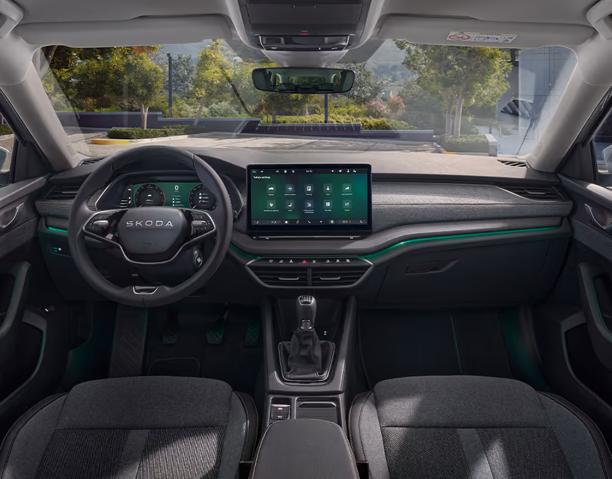 Eine weite Innenansicht zeigt das gesamte Cockpit eines Škoda Octavia mit der Design Selection Lodge. Das Interieur ist mit einer Wollstoff-Kunstleder-Kombination mit gelben Kontrastnähten und einer Dekorleiste in Silber-Grau am Armaturenbrett ausgestattet. Durch die Windschutzscheibe ist eine begrünte, städtische Umgebung zu sehen.