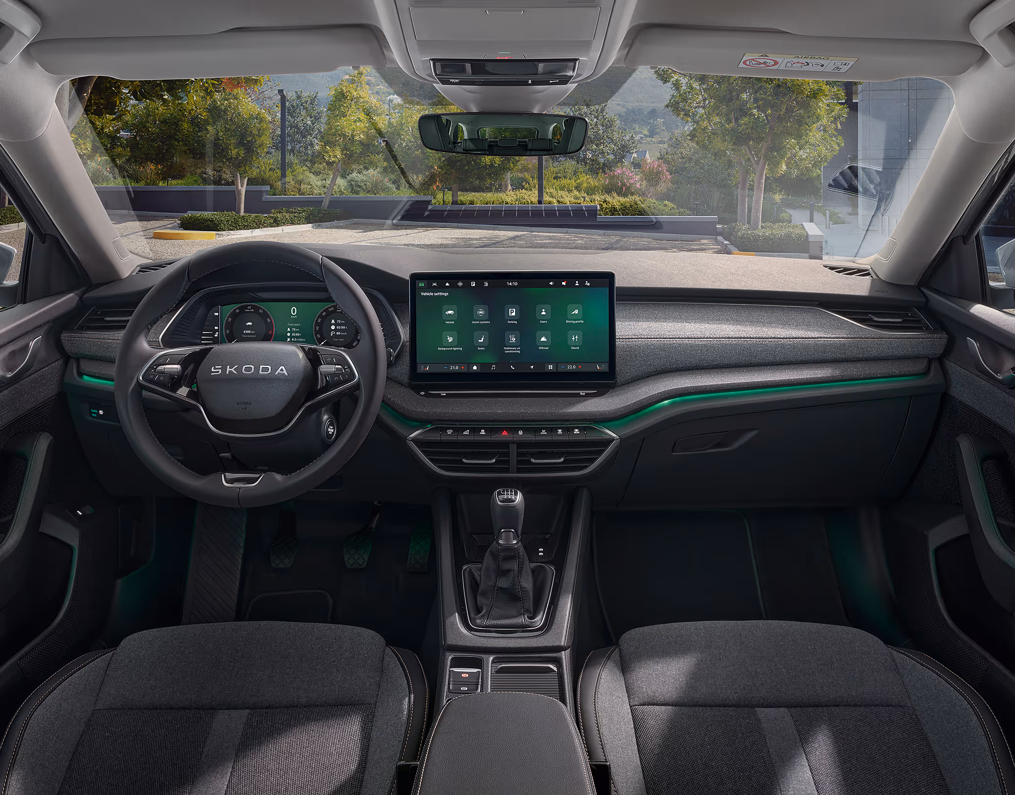 Eine weite Innenansicht zeigt das gesamte Cockpit eines Škoda Octavia mit der Design Selection Lodge. Das Interieur ist mit einer Wollstoff-Kunstleder-Kombination mit gelben Kontrastnähten und einer Dekorleiste in Silber-Grau am Armaturenbrett ausgestattet. Durch die Windschutzscheibe ist eine begrünte, städtische Umgebung zu sehen.