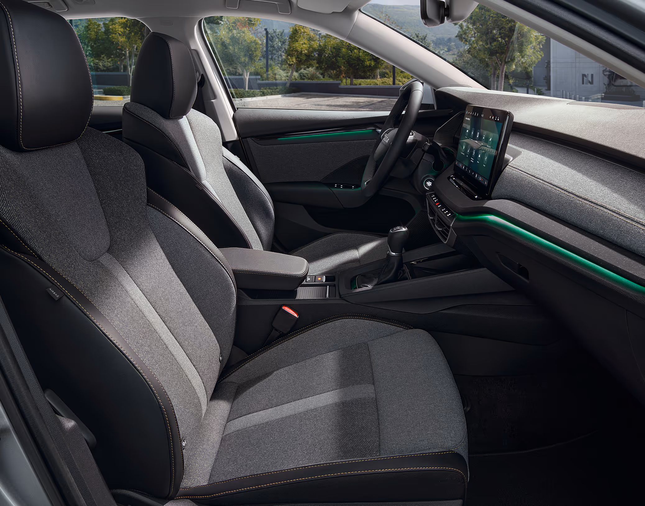 Eine seitliche Innenansicht des Škoda Octavia zeigt die Vordersitze und das Cockpit mit der Design Selection Lodge. Die Sitze aus einer Wollstoff-Kunstleder-Kombination mit gelben Kontrastnähten und die Dekorleiste in Silber-Grau sind zu sehen, während durch die Fenster eine begrünte Außenumgebung sichtbar ist.