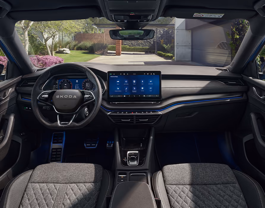 Eine weite Innenansicht zeigt das gesamte Cockpit eines Škoda Octavia mit der Design Selection Sportline. Das sportliche, dunkle Interieur ist mit Sportsitzen aus ThermoFlux-Stoff mit integrierten Kopfstützen und einer Dekorleiste in Carbon-Optik am Armaturenbrett ausgestattet. Durch die Windschutzscheibe ist eine begrünte, städtische Umgebung zu sehen.