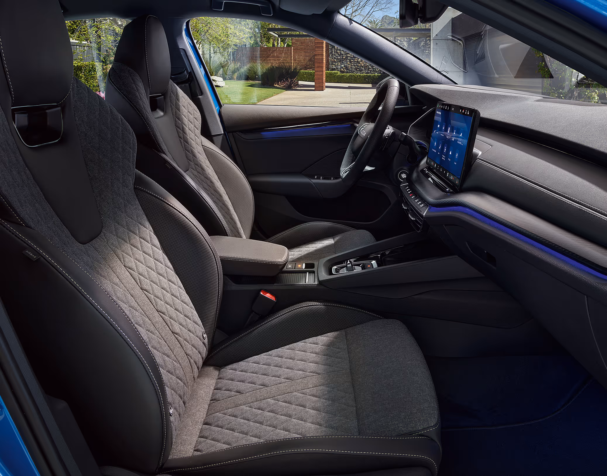 Eine seitliche Innenansicht des Škoda Octavia zeigt die Vordersitze und das Cockpit mit der Design Selection Sportline. Die schwarzen Sportsitze aus ThermoFlux-Stoff mit integrierten Kopfstützen und die Dekorleiste in Carbon-Optik sind zu sehen, während durch die Fenster eine begrünte Außenumgebung sichtbar ist.