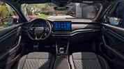 Eine weite Innenansicht zeigt das gesamte Cockpit eines Škoda Octavia mit der Design Selection Sportline. Das sportliche, dunkle Interieur ist mit Sportsitzen aus ThermoFlux-Stoff mit integrierten Kopfstützen und einer Dekorleiste in Carbon-Optik am Armaturenbrett ausgestattet. Durch die Windschutzscheibe ist eine begrünte, städtische Umgebung zu sehen.