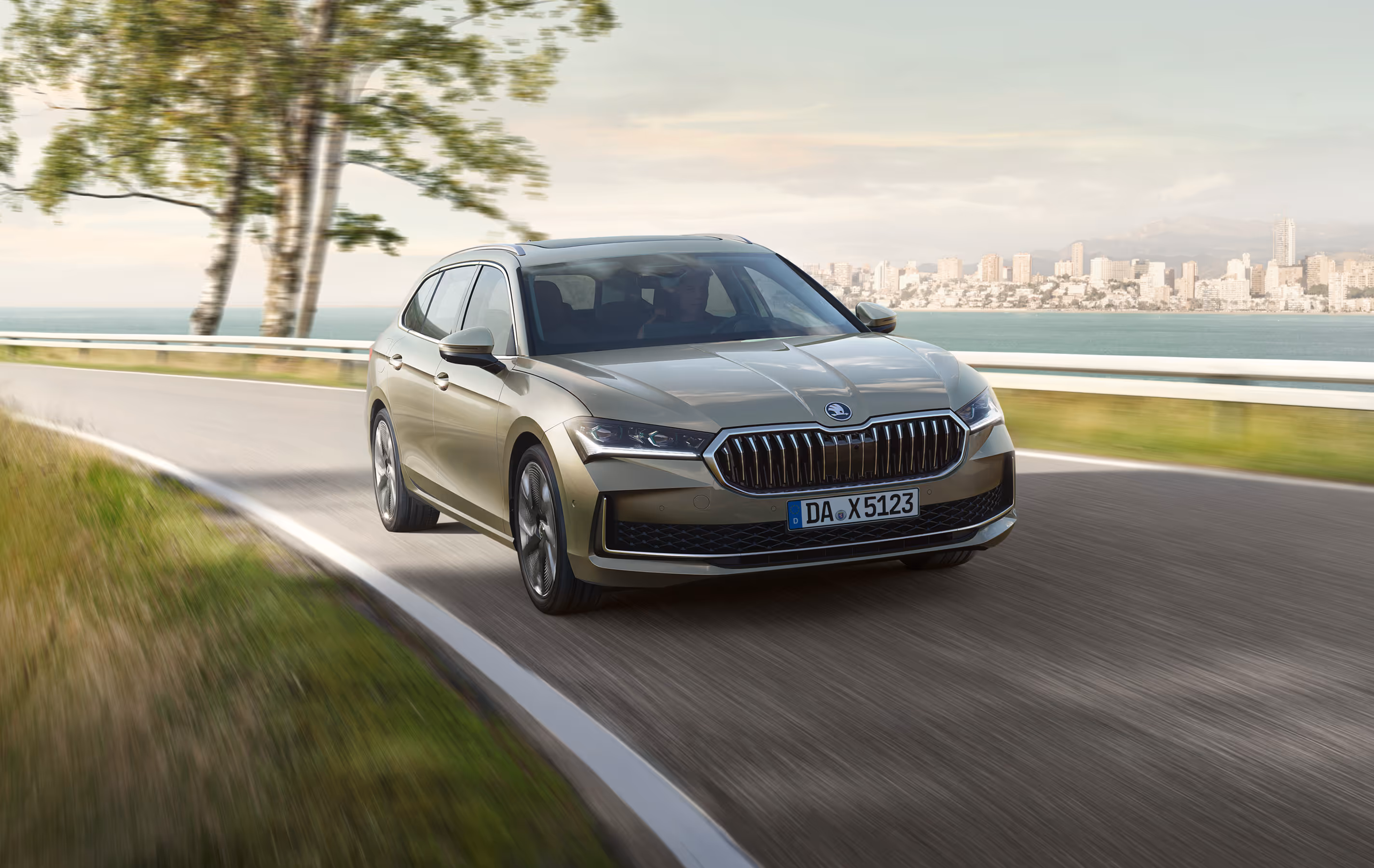 Ein Škoda Superb Combi in der Lackierung Ice-Tea-Beige Metallic fährt auf einer Küstenstraße. Das Fahrzeug ist in einer dynamischen Dreiviertelansicht von vorne abgebildet, während im Hintergrund eine Stadtkulisse über dem Wasser zu sehen ist. 