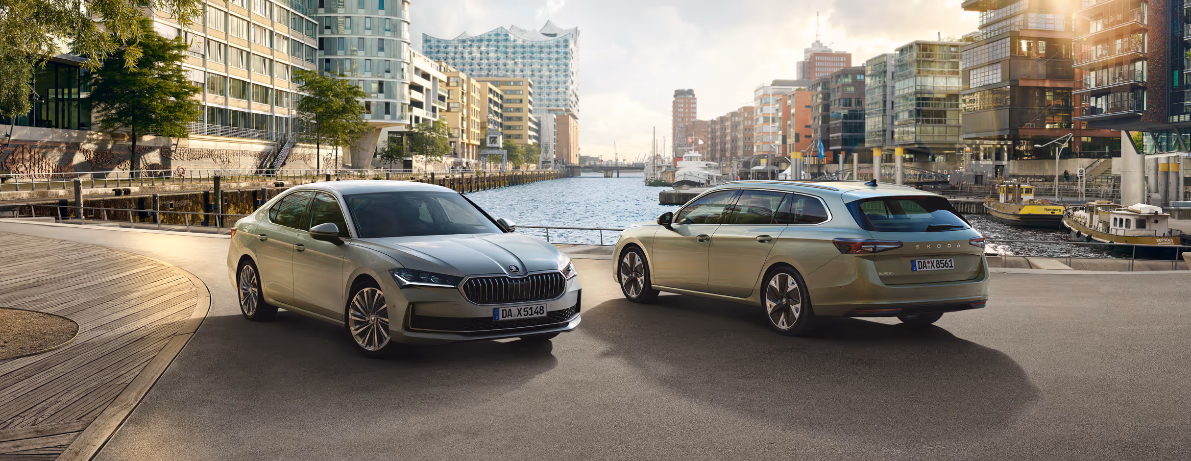 Zwei Škoda Superb Modelle, eine Limousine und ein Combi, sind an einer Hafenpromenade geparkt. Die Limousine in Aluminium-Silber Metallic wird von vorne gezeigt, der Combi in Ice-Tea-Beige Metallic von schräg hinten. Im Hintergrund sind moderne Architektur, Schiffe und die Hamburger Elbphilharmonie bei sonnigem Wetter zu sehen.