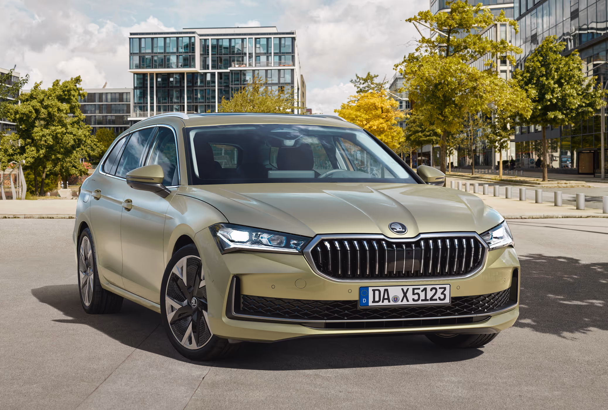 Ein Škoda Superb Combi in der Lackierung Ice-Tea-Beige Metallic ist auf einem Platz in einer städtischen Umgebung geparkt. Das Fahrzeug wird in einer Dreiviertelansicht von vorne gezeigt. Im Hintergrund sind moderne Bürogebäude und Bäume unter einem leicht bewölkten Himmel zu sehen.
