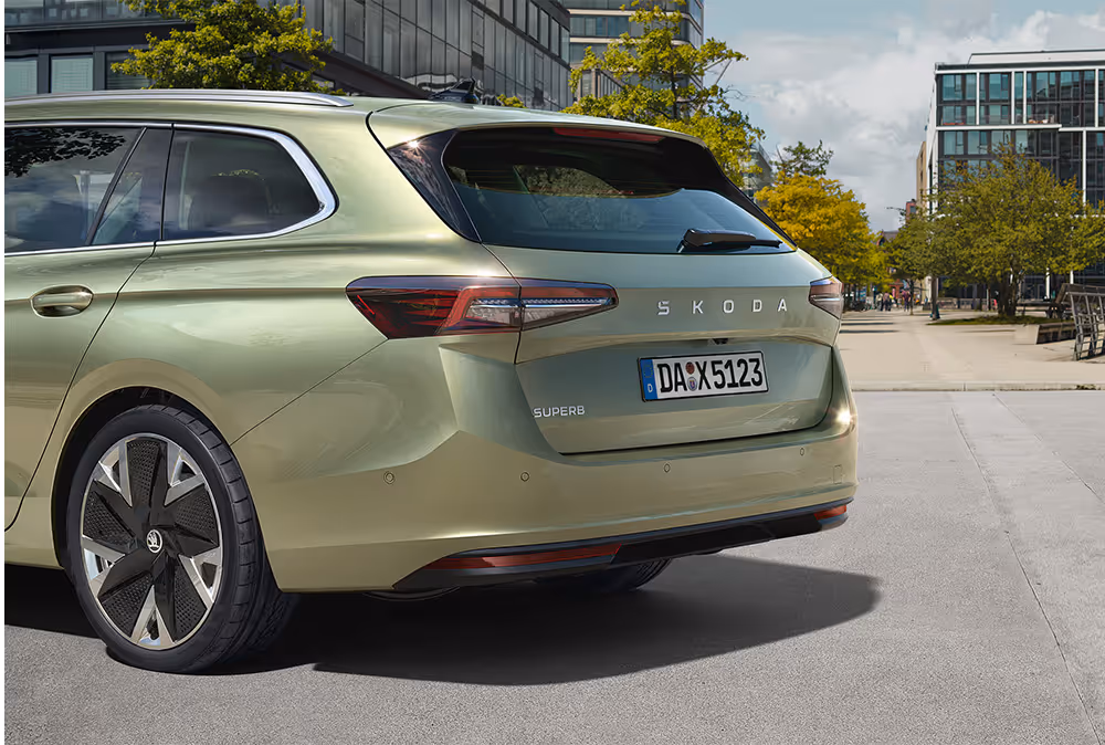 Ein Škoda Superb Combi in der Lackierung Ice-Tea-Beige Metallic ist auf einem städtischen Platz geparkt. Die Aufnahme von schräg hinten hebt das Heckdesign mit den schmalen LED-Heckleuchten und dem neuen Škoda Schriftzug auf der Heckklappe hervor. Im Hintergrund sind moderne Gebäude und eine von Bäumen gesäumte Straße zu erkennen.