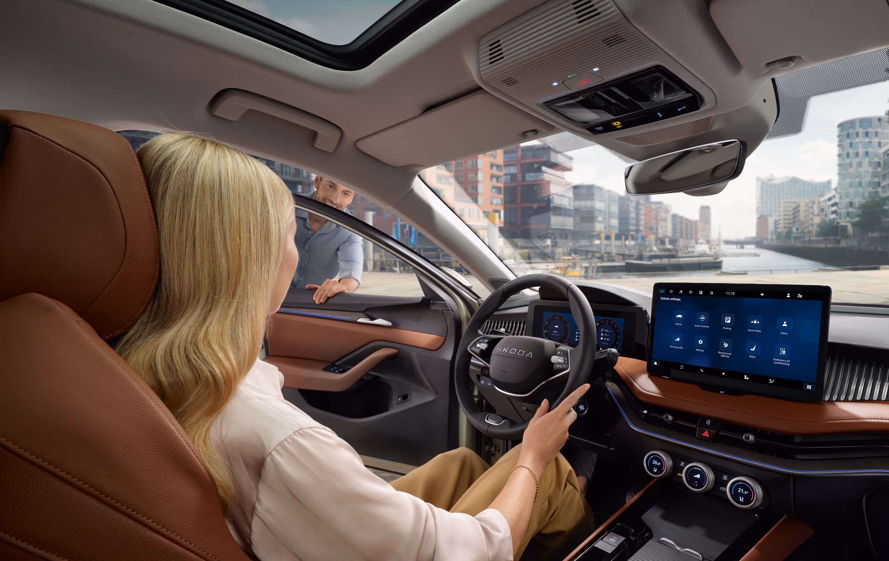Eine Frau sitzt auf dem Fahrersitz eines Škoda Superb und unterhält sich mit einem Mann, der sich durch die geöffnete Fahrertür lehnt. Die Innenansicht zeigt das Cockpit mit der Design Selection Suite in Cognac, dem großen Infotainmentdisplay und dem digitalen Cockpit. Durch die Windschutzscheibe ist die Hamburger HafenCity mit der Elbphilharmonie zu sehen.