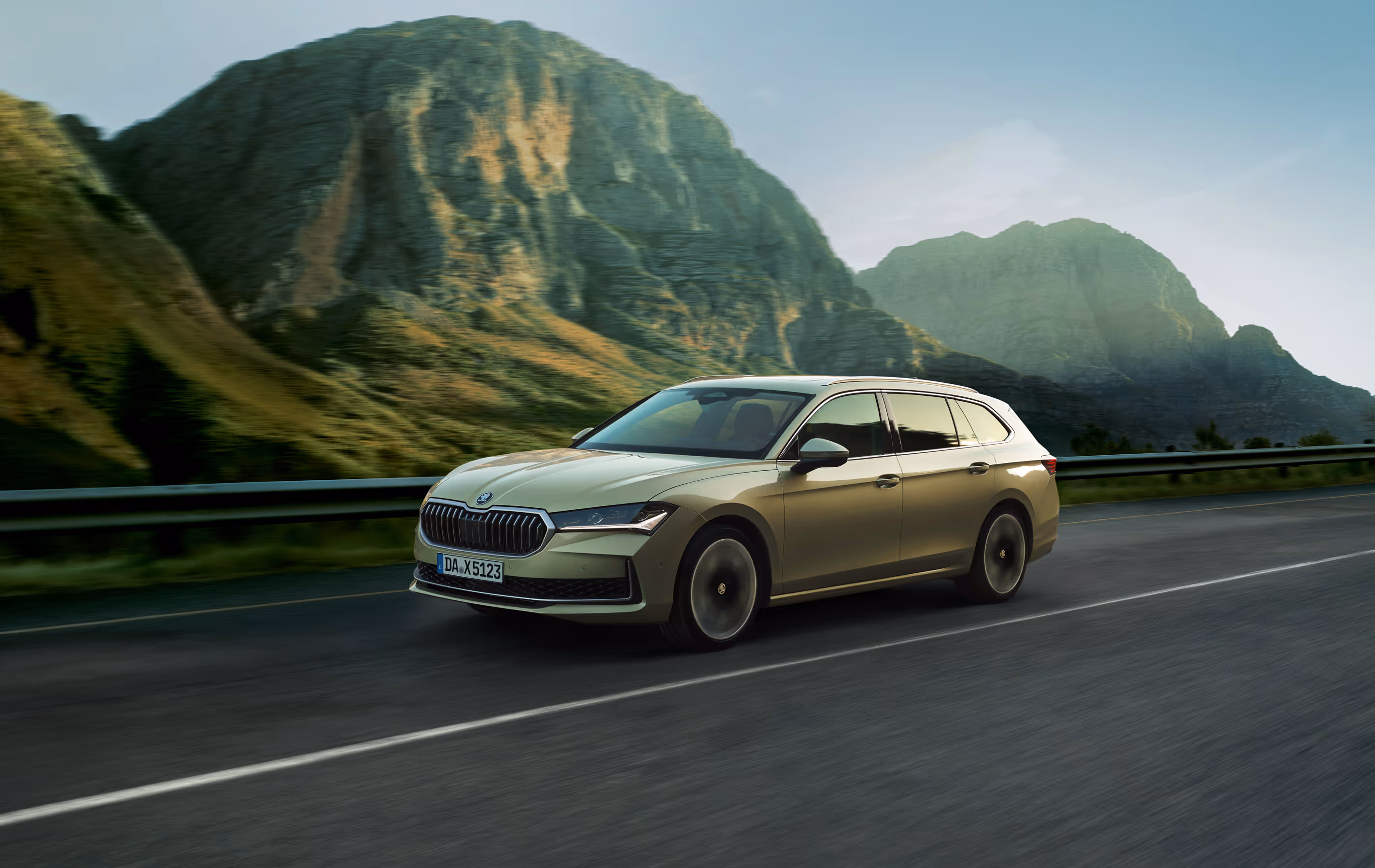 Ein Škoda Superb Combi in der Lackierung Ice-Tea-Beige Metallic fährt auf einer leeren Straße durch eine beeindruckende Berglandschaft. Das Fahrzeug wird in einer dynamischen Dreiviertelansicht von vorne gezeigt, wobei die Bewegungsunschärfe des Hintergrunds die Fahrszene unterstreicht.