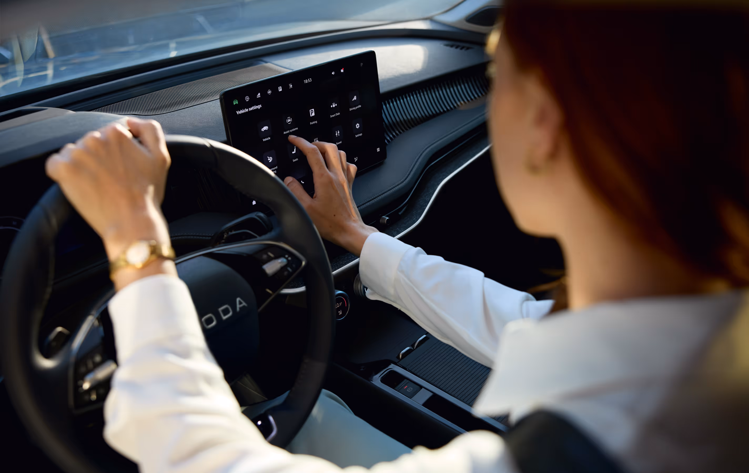 Eine Ansicht über die Schulter einer Fahrerin zeigt, wie sie das Menü für die Fahrzeugeinstellungen auf dem großen, freistehenden Infotainmentdisplay eines Škoda Superb bedient. Während ihre linke Hand am Multifunktionslenkrad liegt, berührt die rechte Hand den Touchscreen. Im modernen Fahrzeuginnenraum sind zudem das digitale Cockpit hinter dem Lenkrad und die multifunktionalen Smart Dials in der Mittelkonsole zu erkennen.

