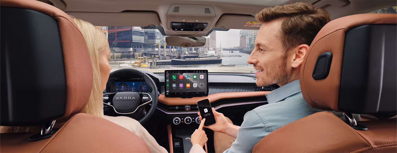 Eine Ansicht aus dem Innenraum eines Škoda Superb zeigt die Nutzung der Wireless SmartLink Funktion. Während eine Frau am Steuer sitzt, hält der Beifahrer ein Smartphone, dessen Benutzeroberfläche über Apple CarPlay auf dem großen, freistehenden Infotainmentdisplay gespiegelt wird. Die Szene spielt sich vor der Kulisse der Hamburger HafenCity ab und zeigt das Interieur der Design Selection Suite in Cognac.
