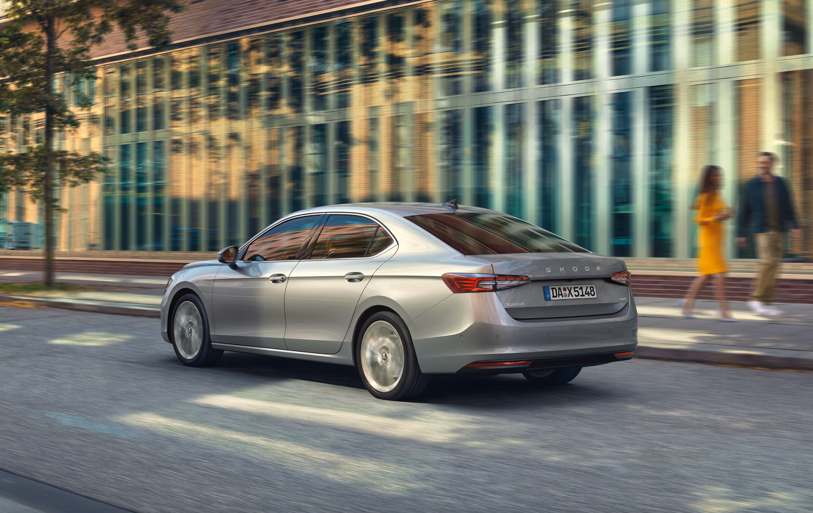 Eine Škoda Superb Limousine in der Lackierung Aluminium-Silber Metallic fährt auf einer Straße durch eine städtische Umgebung. Die dynamische Aufnahme von schräg hinten zeigt das Fahrzeug in Bewegung, was durch den unscharfen Hintergrund mit modernen Gebäuden und Passanten unterstrichen wird.