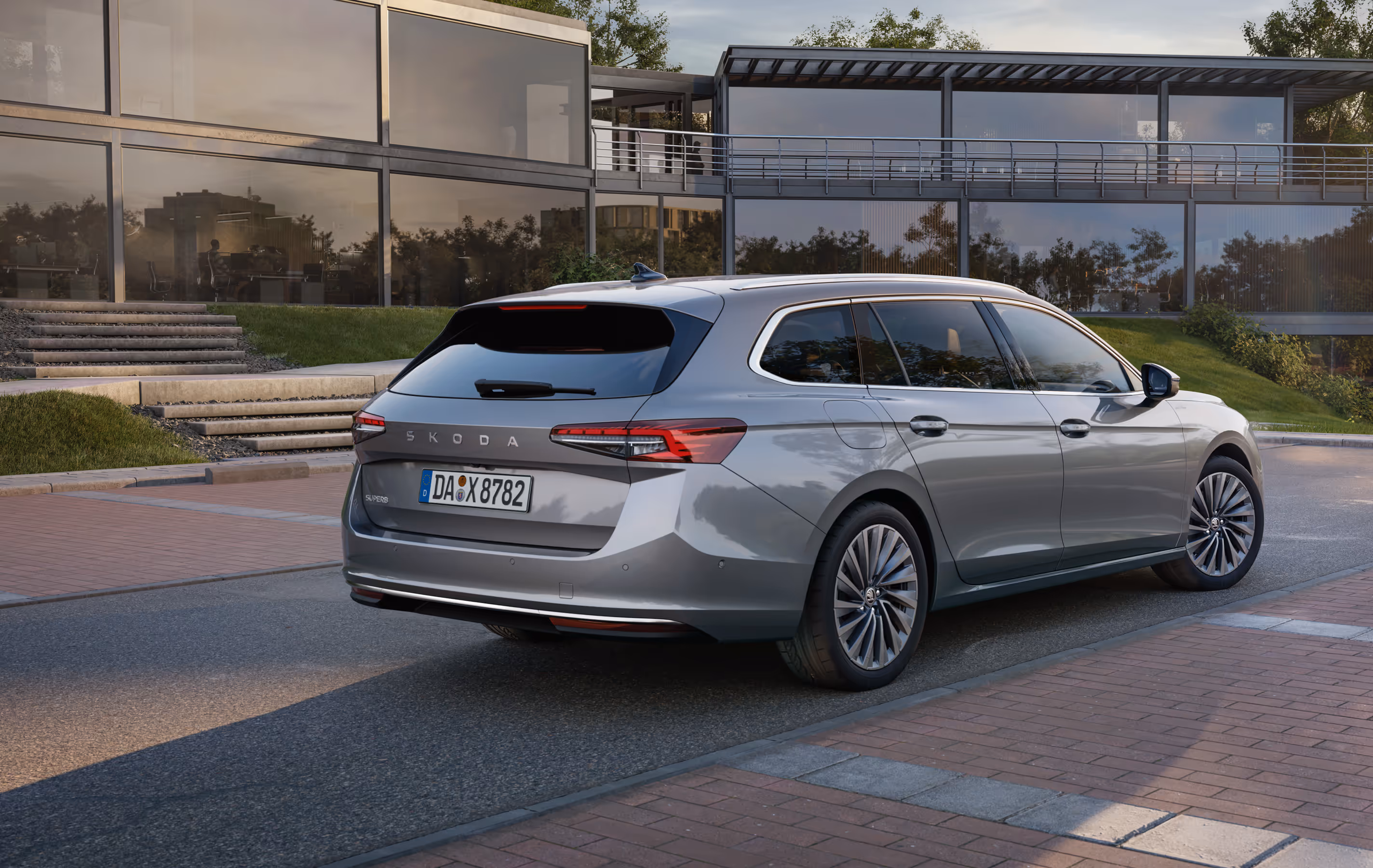 Ein Škoda Superb Combi in der Lackierung Aluminium-Silber Metallic ist auf einem gepflasterten Vorplatz vor einem modernen Bürogebäude geparkt. Die Aufnahme von schräg hinten zeigt die gestreckte Seitenlinie und das Heck des Fahrzeugs mit seinen schmalen LED-Heckleuchten.