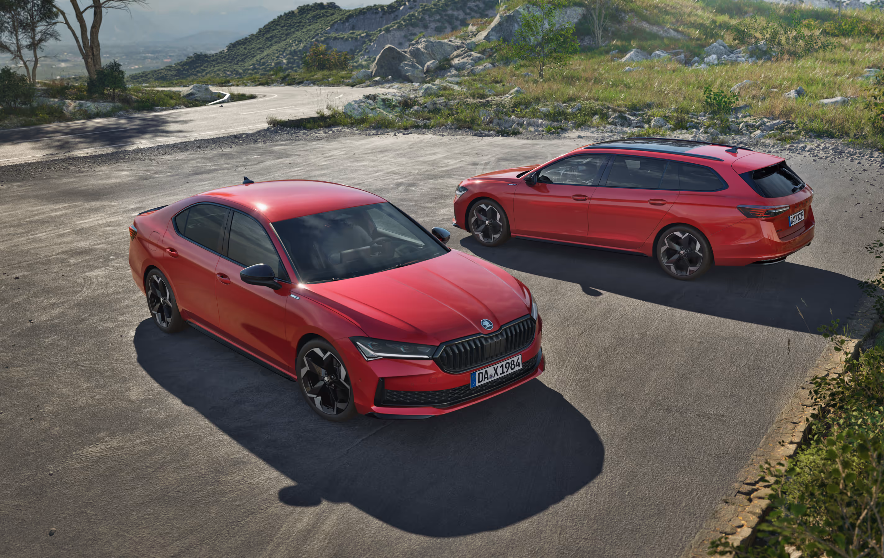 Zwei Škoda Superb Sportline Modelle, eine Limousine und ein Combi, in der Lackierung Karmin-Rot Metallic sind auf einer asphaltierten Fläche in einer bergigen Landschaft geparkt. Die Limousine steht im Vordergrund und wird aus einer erhöhten Dreiviertelperspektive von vorne gezeigt, während der Combi dahinter positioniert ist. 
