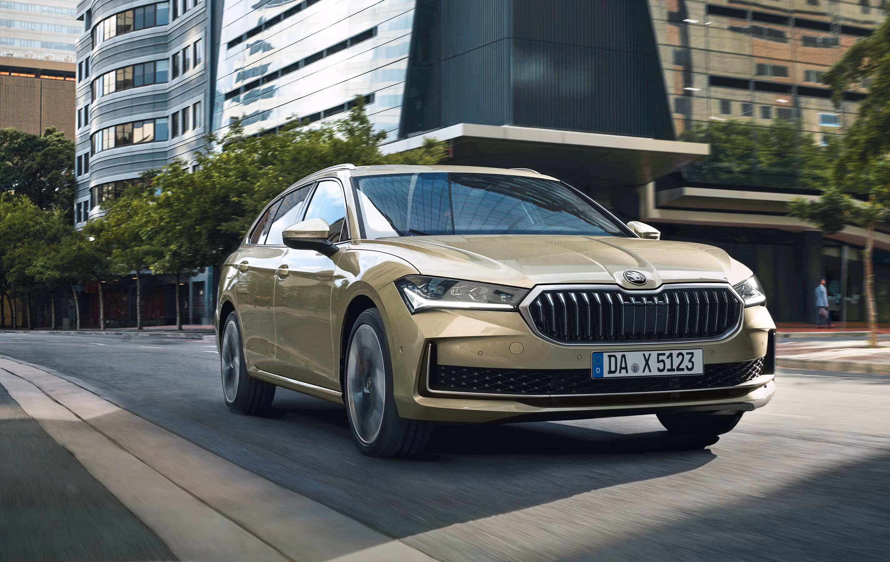 Ein Škoda Superb Combi in der Lackierung Ice-Tea-Beige Metallic fährt auf einer Straße durch eine moderne Stadt. Das Fahrzeug wird in einer dynamischen Dreiviertelansicht von vorne gezeigt, wobei die Bewegungsunschärfe des Hintergrunds mit den Bürogebäuden die Fahrszene unterstreicht.