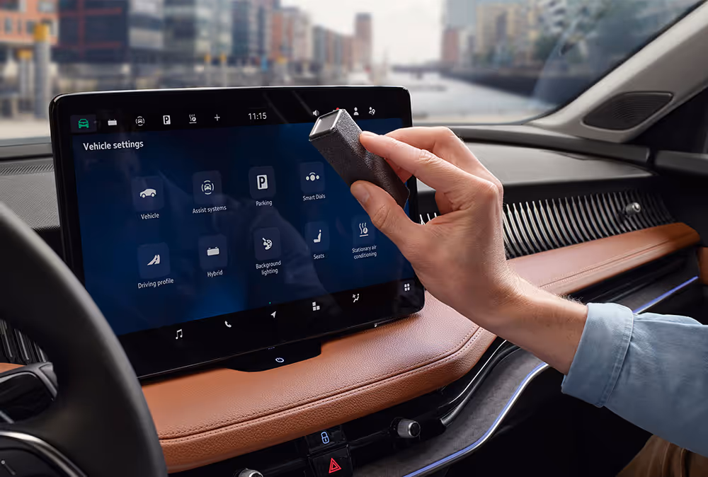 Eine Nahaufnahme zeigt, wie eine Person das große, freistehende Infotainmentdisplay im Cockpit eines Škoda Superb reinigt. Mit dem dafür vorgesehenen Display-Reiniger wird der Bildschirm gesäubert, auf dem gerade das Menü für die Fahrzeugeinstellungen zu sehen ist. Durch die Windschutzscheibe ist die Hamburger HafenCity zu erkennen.