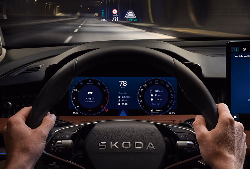 Das Bild zeigt die Fahrerperspektive in einem Škoda Superb während einer Fahrt durch einen Tunnel. Im direkten Sichtfeld des Fahrers projiziert das Head-up-Display wichtige Fahrinformationen wie Geschwindigkeit, Tempolimit und Navigationshinweise auf die Windschutzscheibe. Darunter sind die Hände des Fahrers am Lenkrad und das digitale Cockpit zu sehen, das ebenfalls detaillierte Fahrdaten anzeigt.