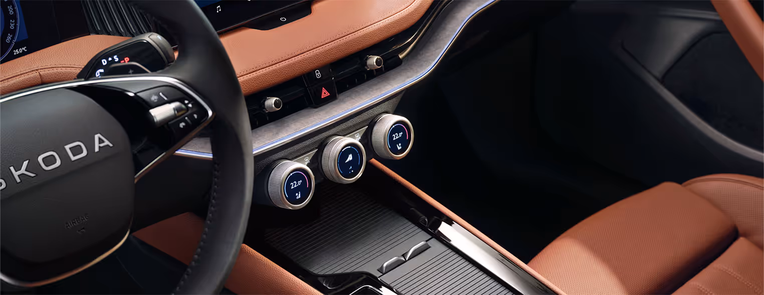 Eine Nahaufnahme der Mittelkonsole eines Škoda Superb zeigt die drei multifunktionalen Smart Dials mit ihren integrierten Digitalanzeigen. Die Konsole ist Teil der Design Selection Suite in Cognac und von cognac-farbenem Leder sowie einer dunklen Dekorleiste umgeben. Links im Bild sind ein Teil des Lenkrads und der Wählhebel an der Lenksäule zu erkennen.