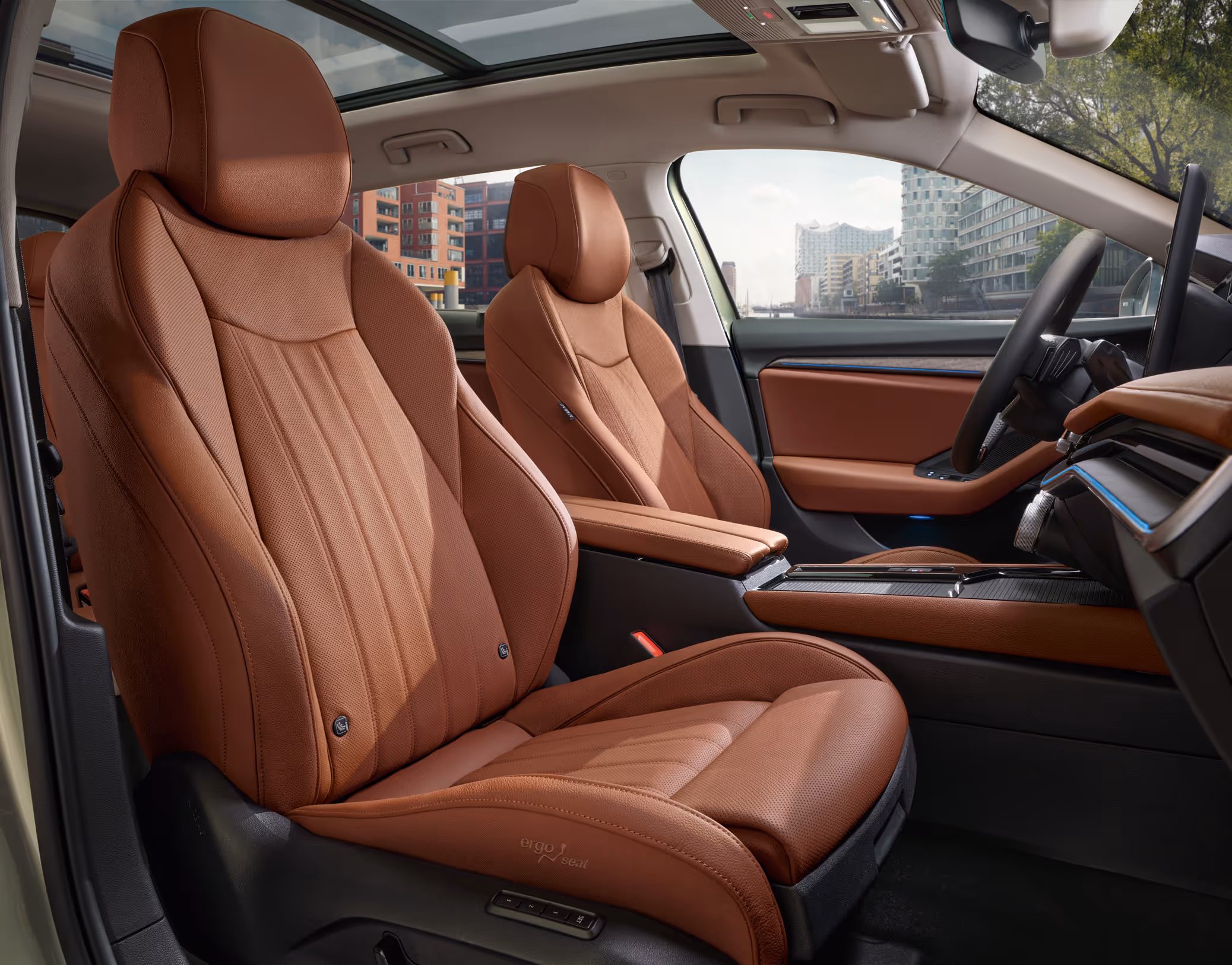 Eine seitliche Innenansicht des Škoda Superb zeigt die vorderen Ergo-Komfortsitze mit Massagefunktion und das Cockpit mit der Design Selection Suite in Cognac. Die cognac-braunen, perforierten Ledersitze werden von oben durch das Panoramaschiebedach beleuchtet.