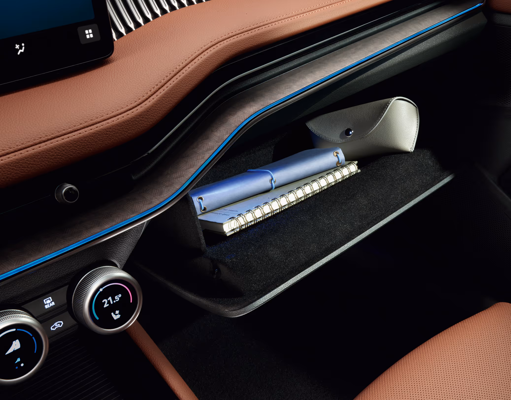 Eine Nahaufnahme zeigt das geöffnete Handschuhfach im Cockpit eines Škoda Superb. Das Fach ist mit einem Notizbuch und einem Brillenetui gefüllt. Das Interieur der Design Selection Suite in Cognac wird durch die blaue Ambientebeleuchtung akzentuiert.