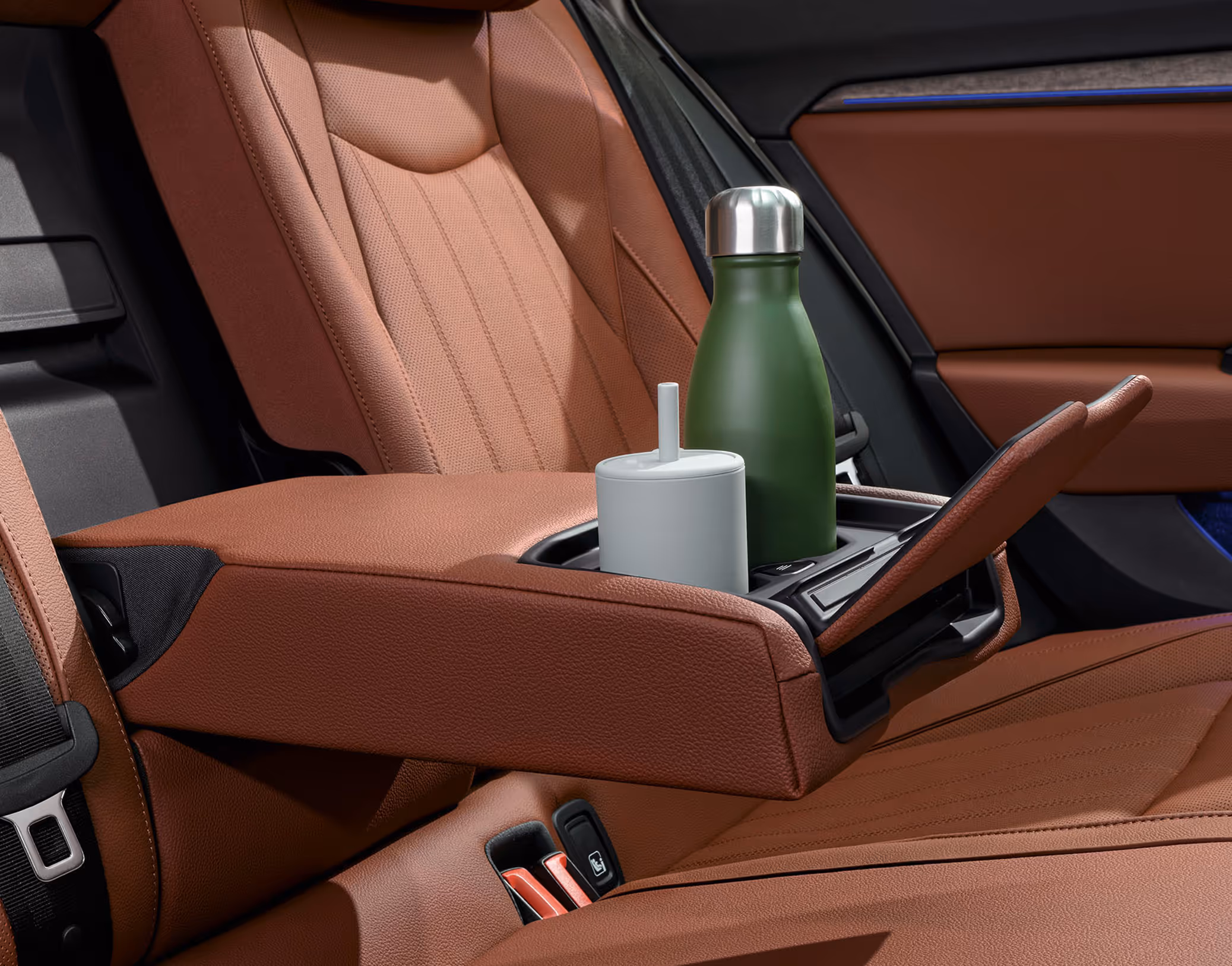 Eine Nahaufnahme der Rücksitzbank eines Škoda Superb zeigt die heruntergeklappte Mittelarmlehne der Design Selection Suite. Die Armlehne aus cognac-farbenem Leder ist mit Getränkehaltern ausgestattet, in denen eine grüne Trinkflasche und ein weißer Becher stehen.
