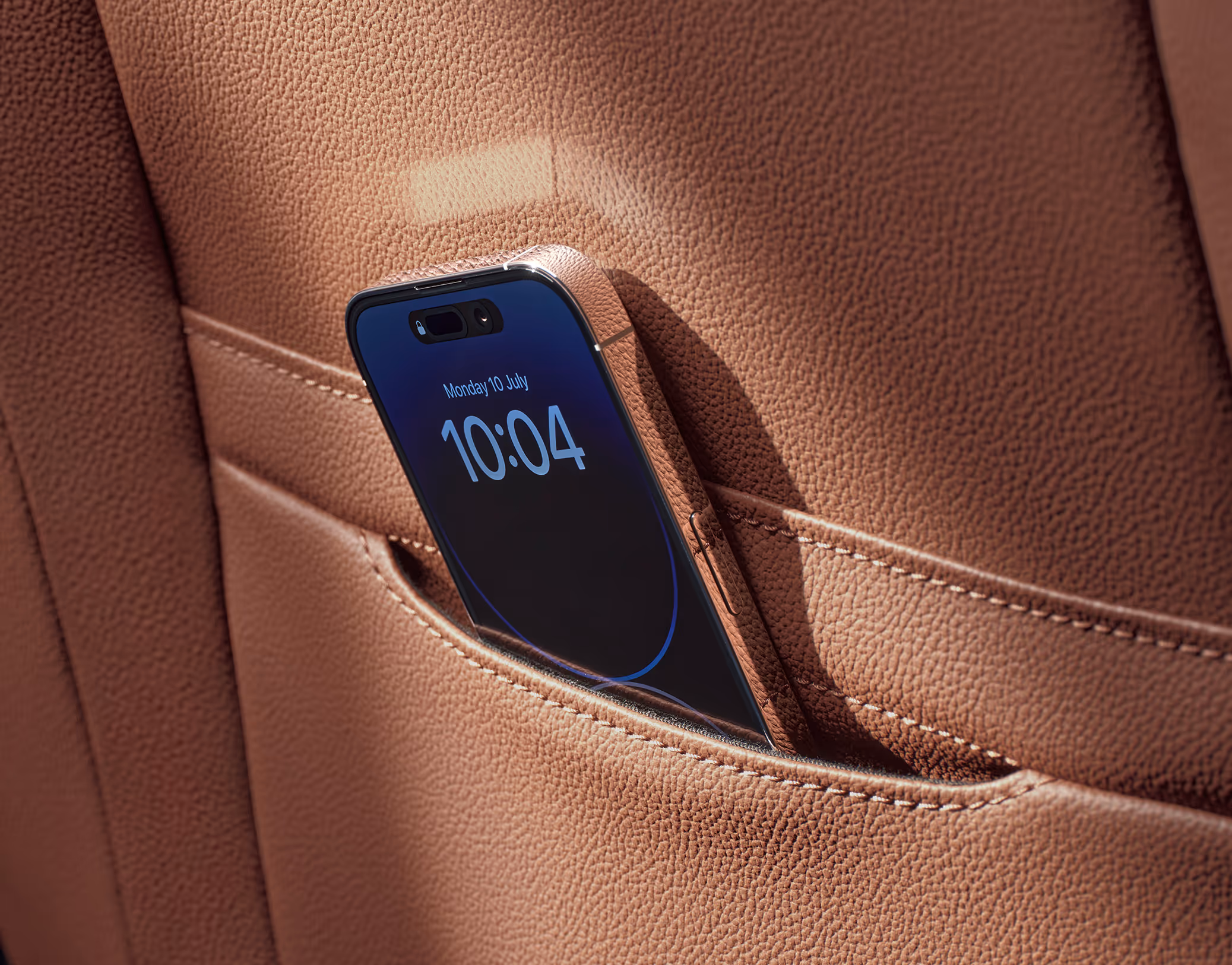 Eine Nahaufnahme zeigt die Smartphone-Tasche an der Rückseite eines Vordersitzes im Škoda Superb. Ein Smartphone mit eingeschaltetem Display, das die Uhrzeit 10:04 anzeigt, steckt in der Tasche des cognac-farbenen Ledersitzes.