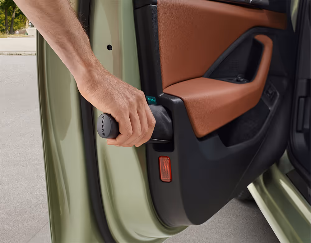 Eine Nahaufnahme zeigt, wie eine Hand den integrierten Regenschirm aus seinem speziellen Fach in der Fahrertür eines Škoda Superb entnimmt. Das Fach ist Teil der Türinnenverkleidung, die mit einer cognac-farbenen Lederarmlehne ausgestattet ist.