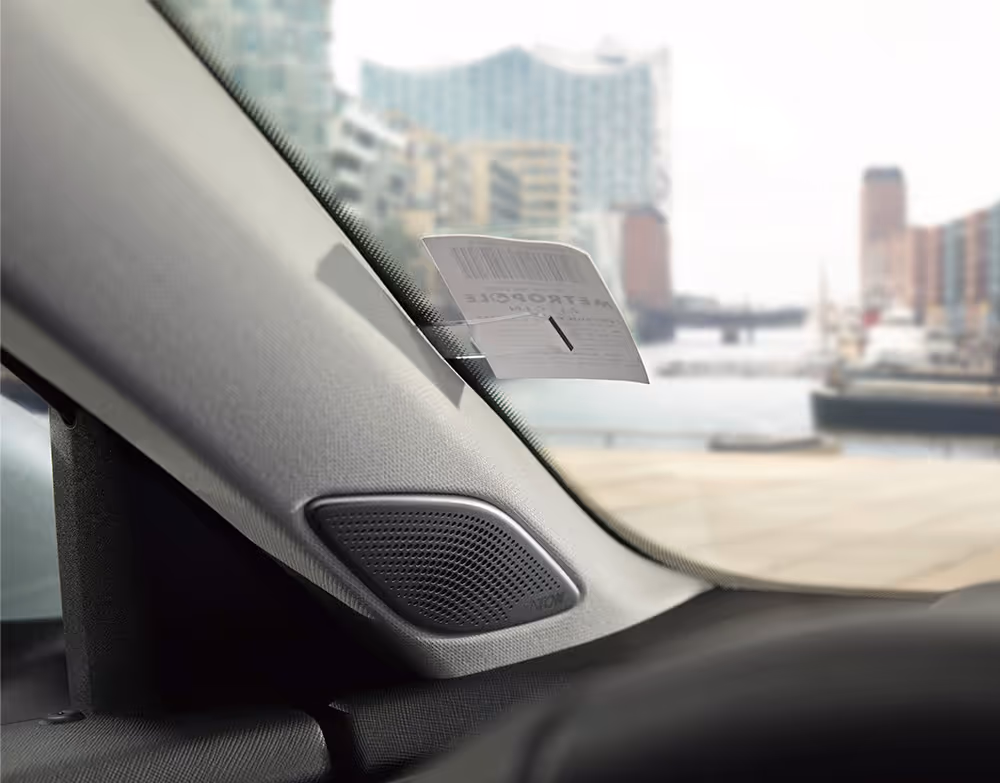 Eine Nahaufnahme aus dem Innenraum eines Škoda Superb zeigt den Tickethalter an der A-Säule, in dem ein Parkschein steckt. Darunter ist der Lautsprecher des Soundsystems in die Säulenverkleidung integriert. Durch die Windschutzscheibe ist in der Unschärfe die Hamburger Elbphilharmonie zu erkennen.