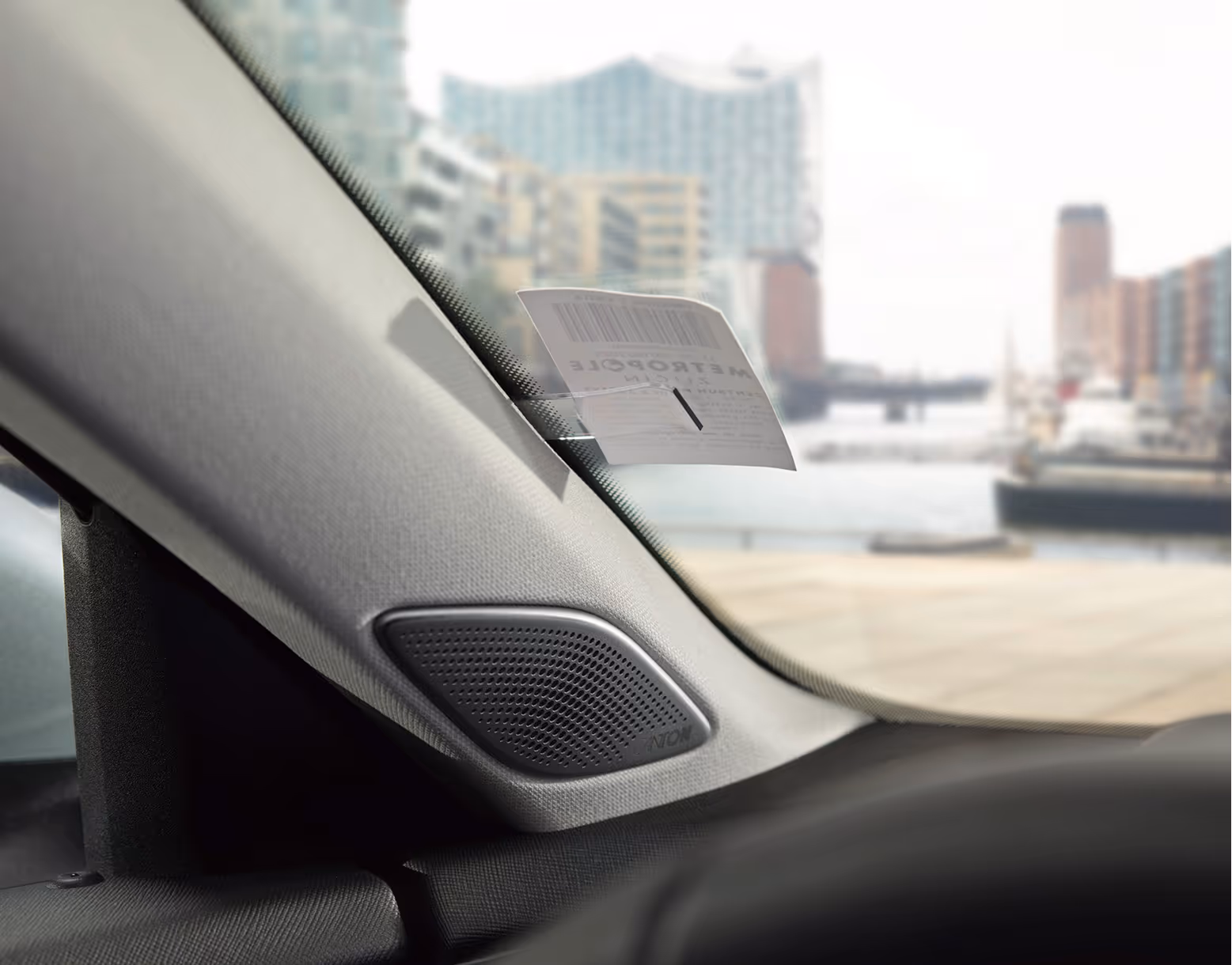 Eine Nahaufnahme aus dem Innenraum eines Škoda Superb zeigt den Tickethalter an der A-Säule, in dem ein Parkschein steckt. Darunter ist der Lautsprecher des Soundsystems in die Säulenverkleidung integriert. Durch die Windschutzscheibe ist in der Unschärfe die Hamburger Elbphilharmonie zu erkennen.