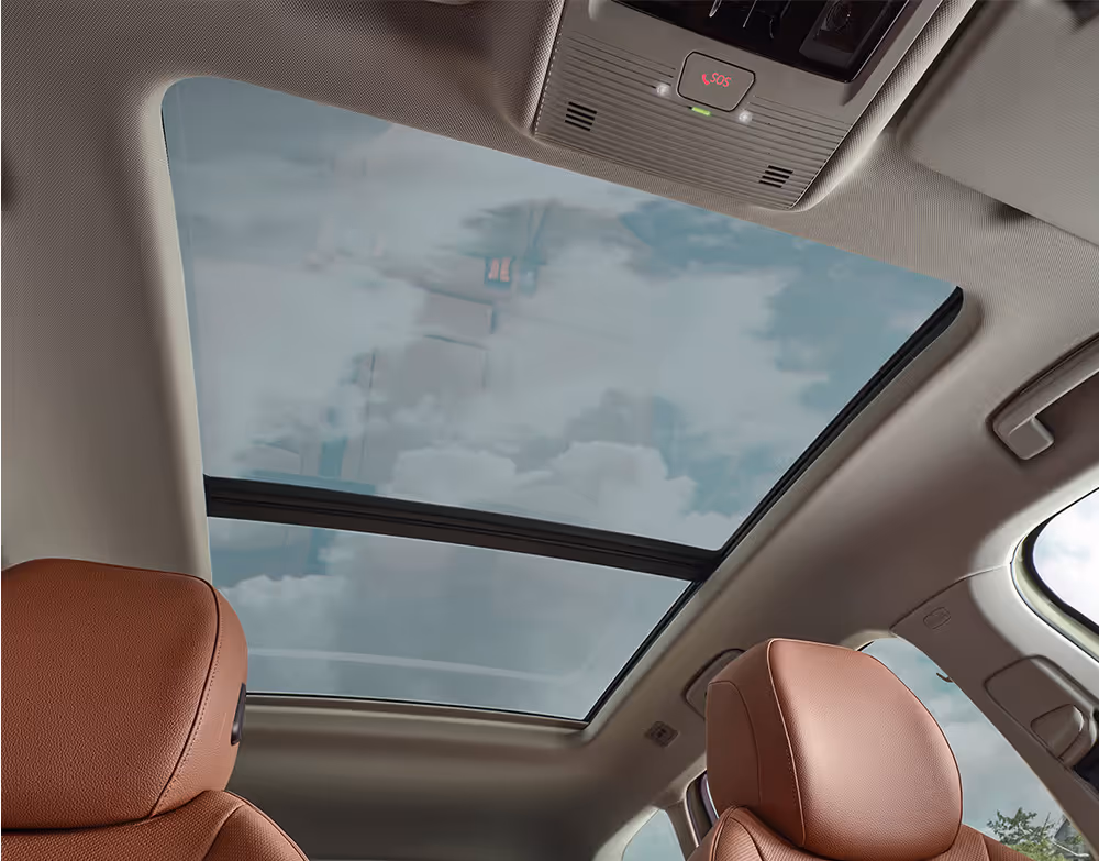 Ein Blick nach oben im Innenraum eines Škoda Superb zeigt das große, optionale Panoramaschiebedach, durch das der leicht bewölkte Himmel zu sehen ist. Die Aufnahme zeigt auch die Kopfstützen der cognac-farbenen Ledersitze und die Dachkonsole mit dem roten SOS-Knopf für den Notruf.