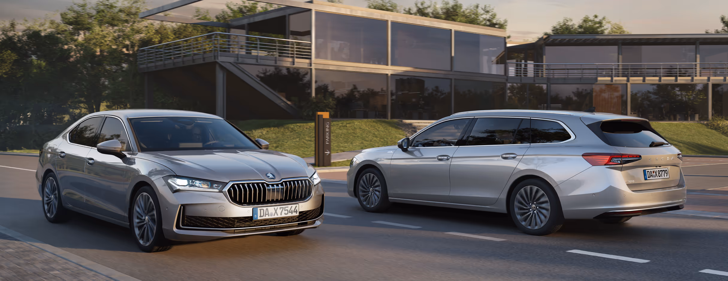 Zwei Škoda Superb Modelle in der Lackierung Aluminium-Silber Metallic, eine Limousine und ein Combi, sind auf einer Straße vor einem modernen Gebäude unterwegs. Die Limousine wird in einer dynamischen Dreiviertelansicht von vorne gezeigt, während der Combi im Hintergrund von schräg hinten zu sehen ist. Die Szene findet bei tiefstehender Sonne statt, die sich in den Glasfassaden des Gebäudes spiegelt.