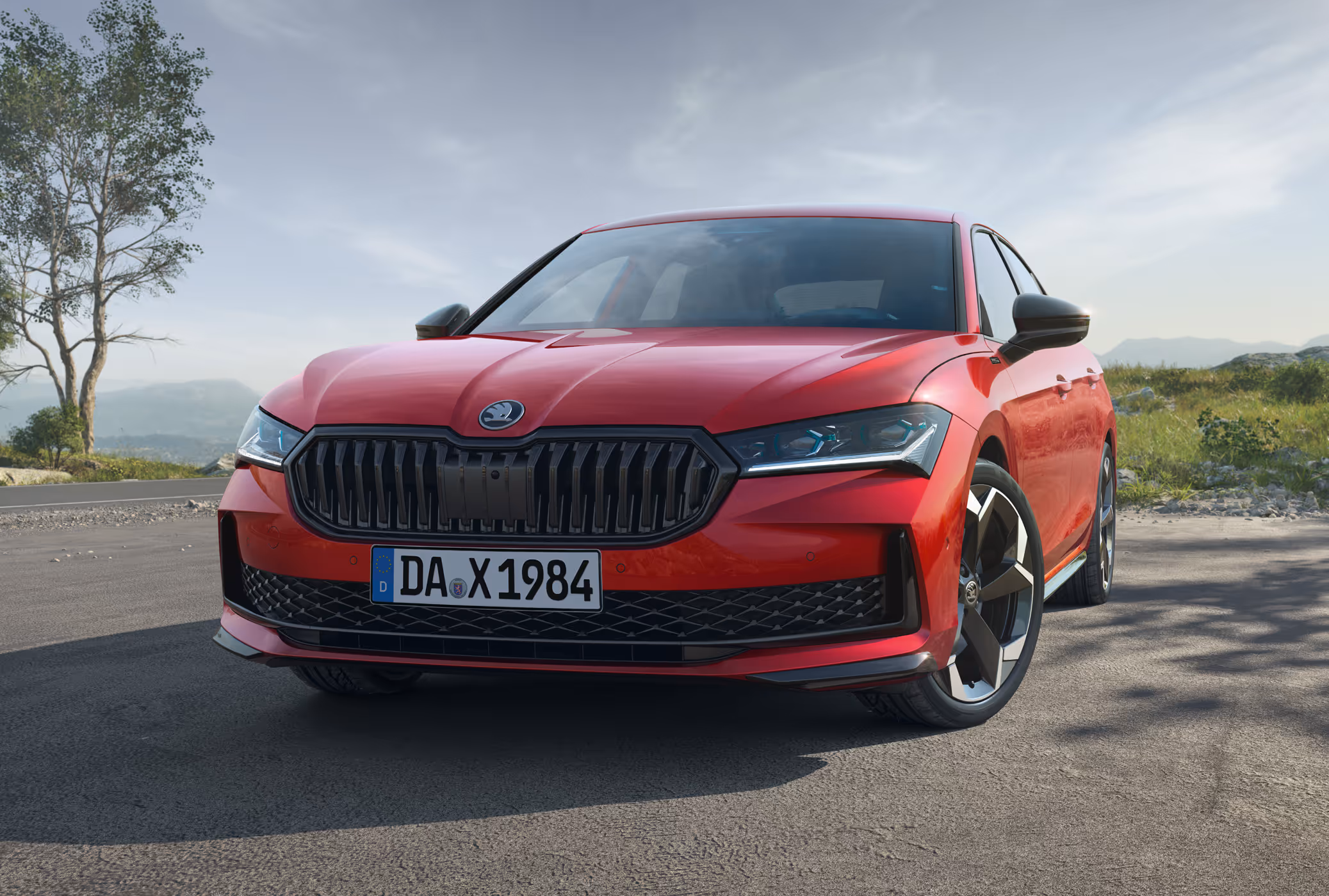 Ein Škoda Superb Sportline Limousine in der Lackierung Karmin-Rot Metallic ist auf einer asphaltierten Straße in einer hügeligen Landschaft geparkt. Die Aufnahme aus einer niedrigen Dreiviertelperspektive von vorne hebt die sportlichen, schwarzen Design-Akzente wie den Kühlergrill und die Außenspiegelkappen hervor. Im Hintergrund sind Bäume und eine Bergkette unter einem leicht bewölkten Himmel zu sehen.
