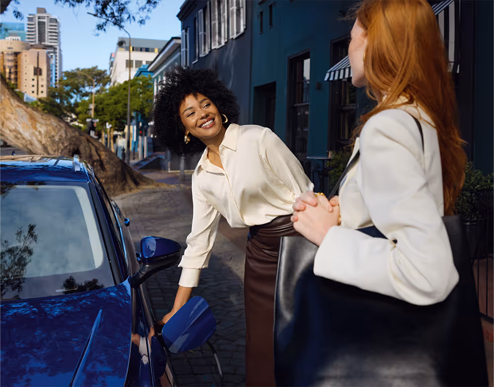 Zwei Frauen unterhalten sich neben einem Škoda Superb Combi iV in der Lackierung Atlantik-Blau Metallic, der an einer Straße in der Stadt lädt. Eine der Frauen lächelt, während sie den Ladestecker am Fahrzeug bedient. Im Hintergrund sind städtische Gebäude und Bäume zu sehen.