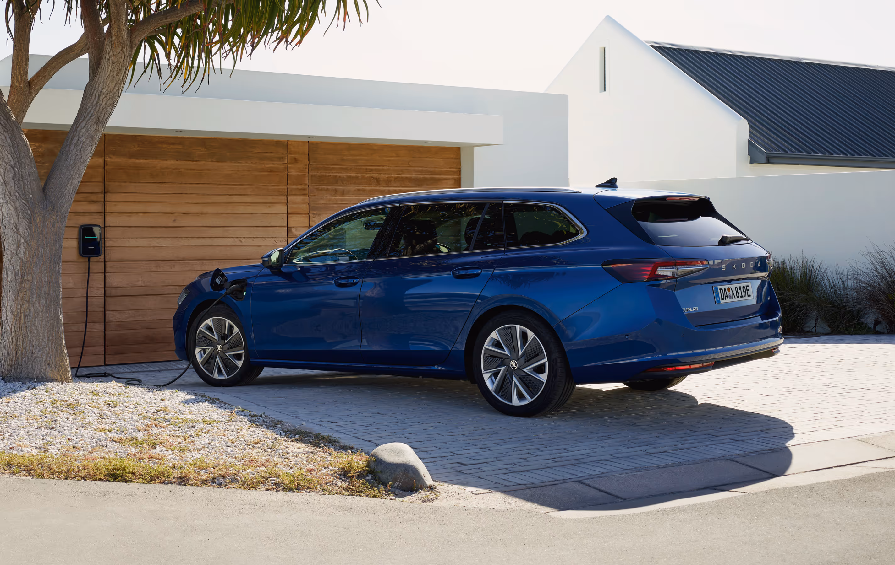 Ein Škoda Superb Combi iV in der Lackierung Atlantik-Blau Metallic lädt an einer Wallbox in der gepflasterten Einfahrt eines modernen Hauses. Das Fahrzeug, das von schräg hinten gezeigt wird, ist über ein Ladekabel mit der an der Holzwand des Hauses montierten Ladestation verbunden.