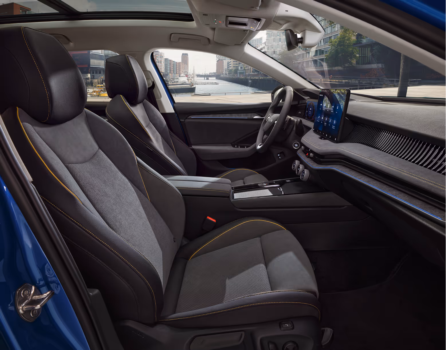 Eine seitliche Innenansicht des Škoda Superb zeigt das Interieur der Design Selection Lounge. Die Sitzbezüge aus einer grauen Suedia-Stoff-Kunstleder-Kombination mit gelben Kontrastnähten sowie das dunkle Armaturenbrett mit einer Dekorleiste in Unique Dark Chrome sind zu sehen, während durch die Fenster die Hamburger HafenCity sichtbar ist.